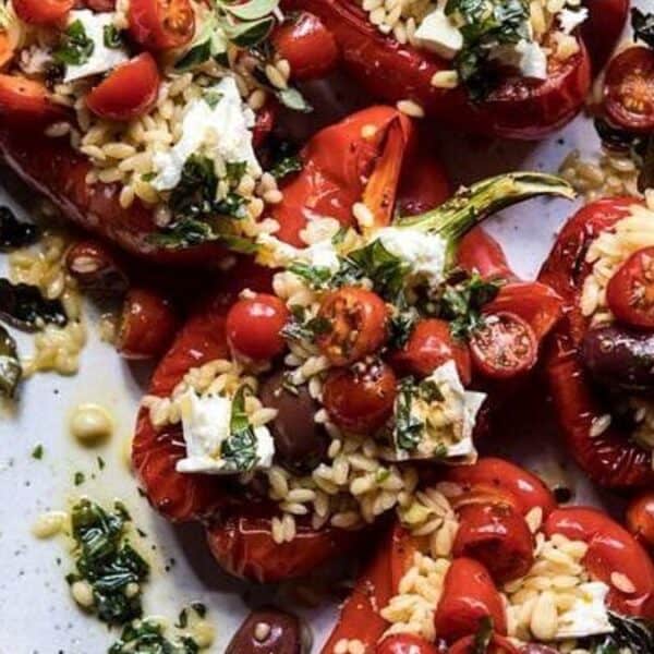 Roasted red bell peppers on a plate, filled with orzo, cherry tomatoes, olives, feta cheese, and topped with fresh herbs.