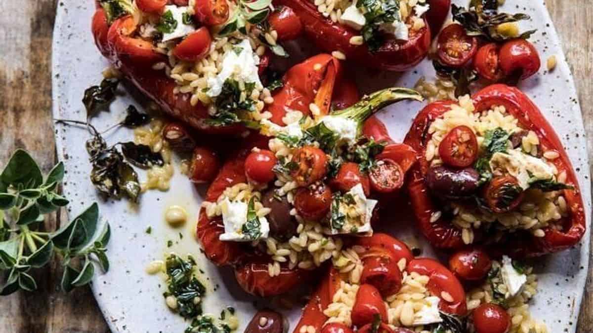 Roasted red bell peppers on a plate, filled with orzo, cherry tomatoes, olives, feta cheese, and topped with fresh herbs.