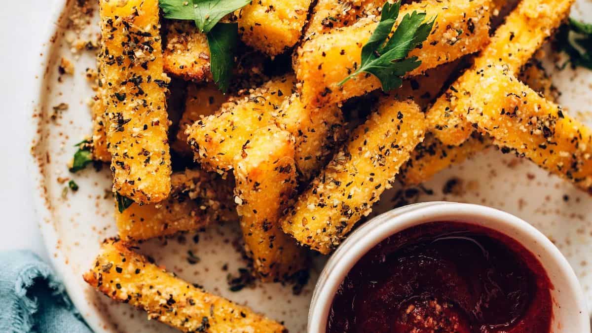 A plate of seasoned polenta fries garnished with herbs, served with a side of red dipping sauce.