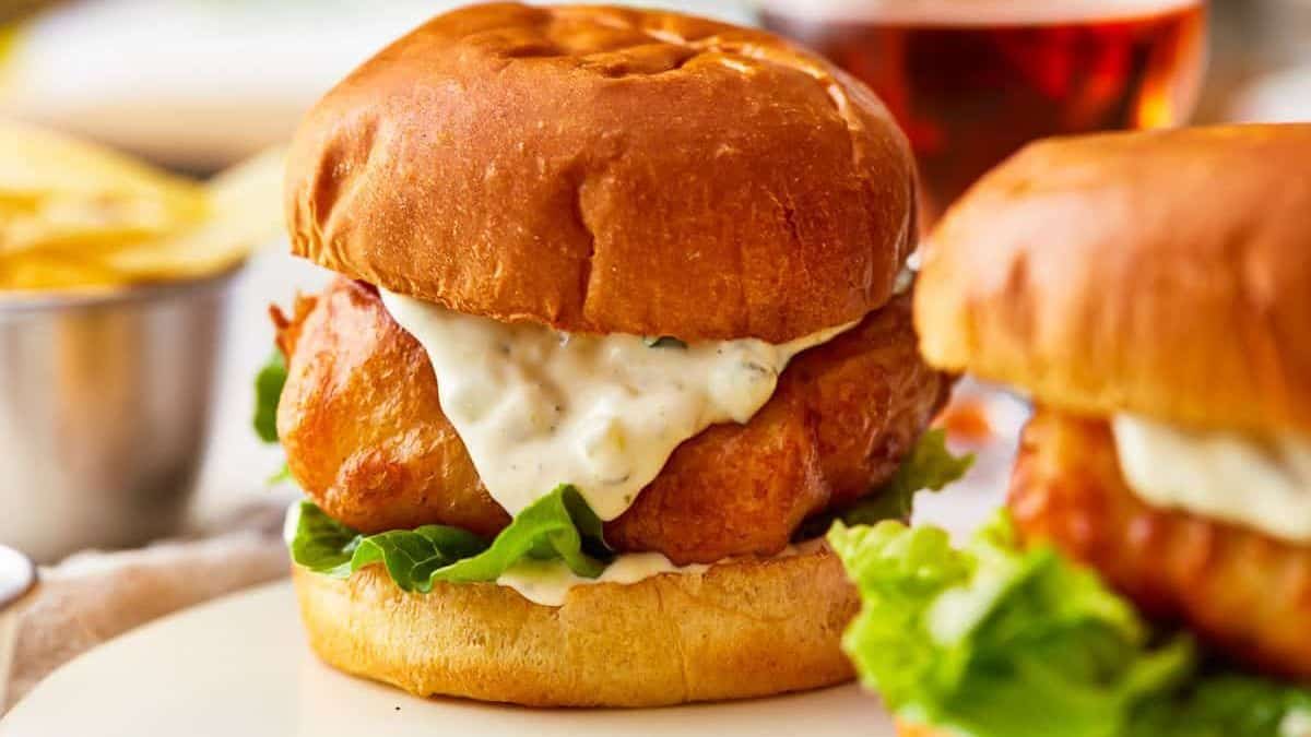 A fried fish sandwich with lettuce and tartar sauce on a toasted bun is on a plate, with chips and a drink behind it.