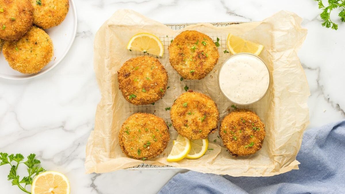Six breaded crab cakes on parchment paper with a bowl of dipping sauce, lemon wedges, and parsley garnish.