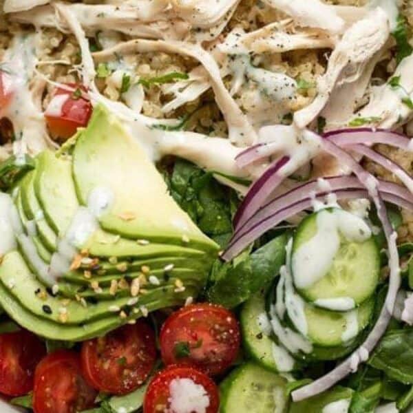 A bowl with shredded chicken, sliced avocado, red onion, cucumber, cherry tomatoes, lettuce, quinoa, and dressing.