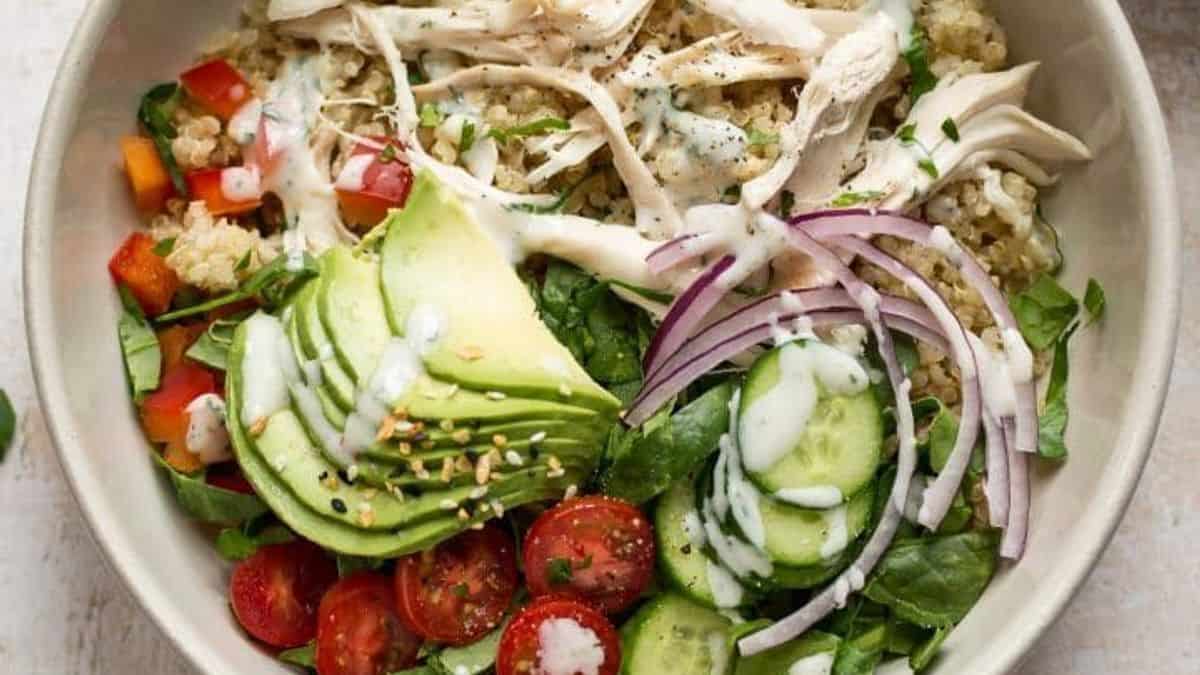 A bowl containing shredded chicken, sliced avocado, cherry tomatoes, cucumber, red onion, peppers, greens, quinoa, and dressing.