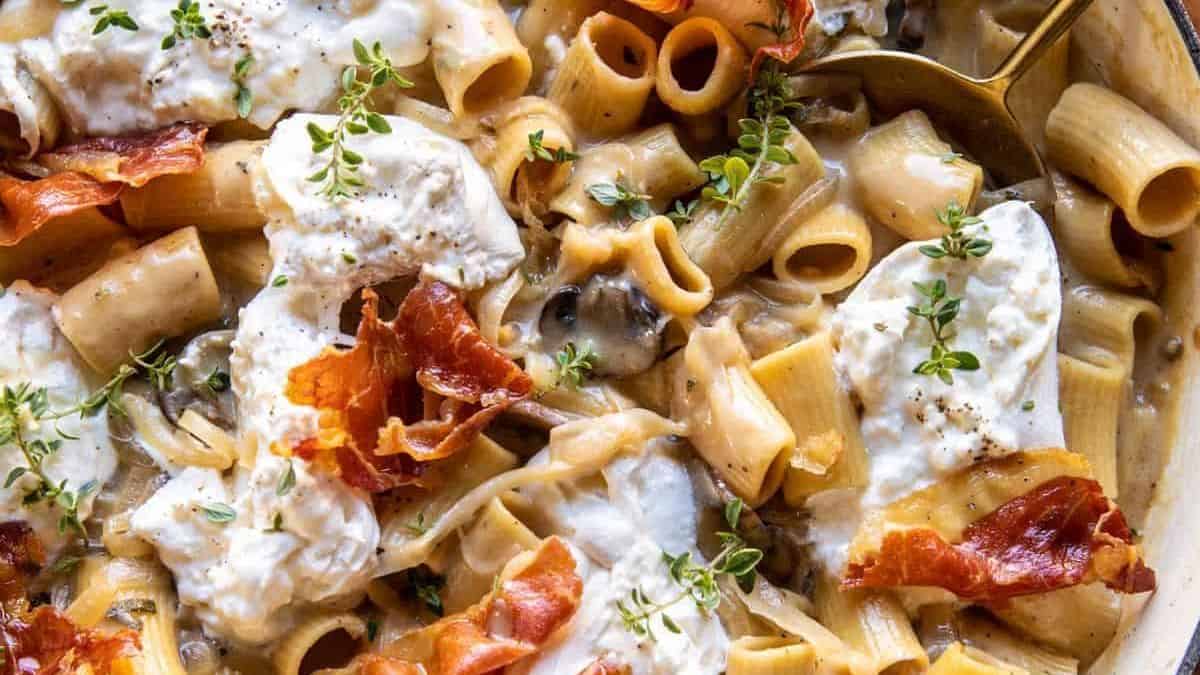 Rigatoni pasta in a creamy sauce with mushrooms, melted cheese, crispy prosciutto, fresh thyme, and a serving spoon.