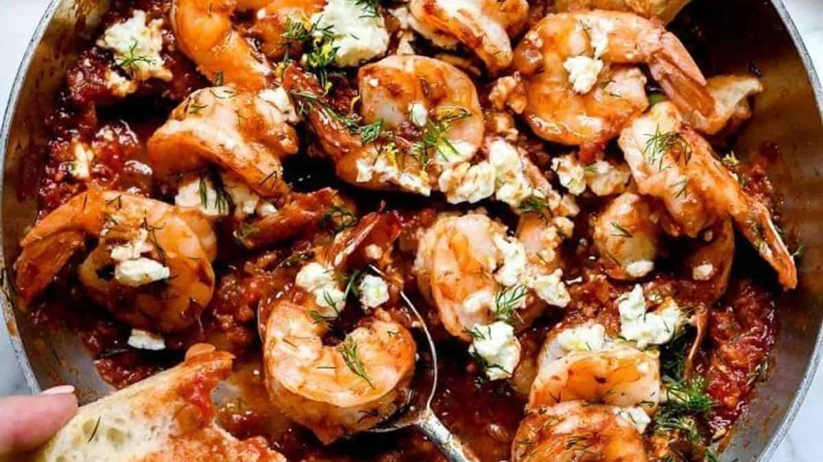 Shrimp in tomato sauce topped with crumbled feta and herbs in a skillet, served with a piece of bread.