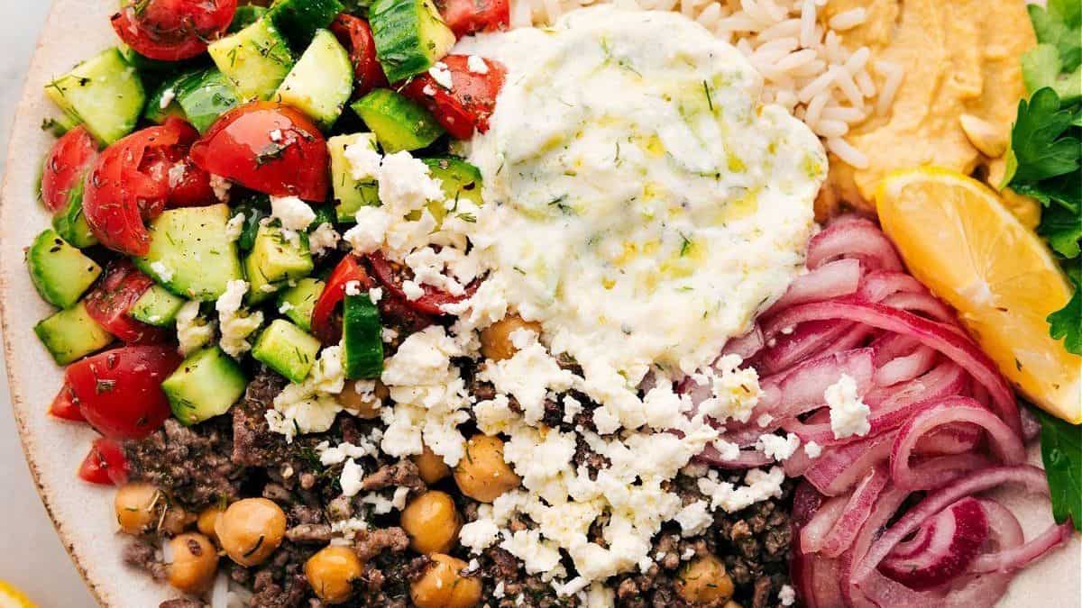 Ground beef, chickpeas, crumbled feta, sliced red onions, cucumber tomato salad, tzatziki, hummus, rice, and a lemon wedge in a bowl.