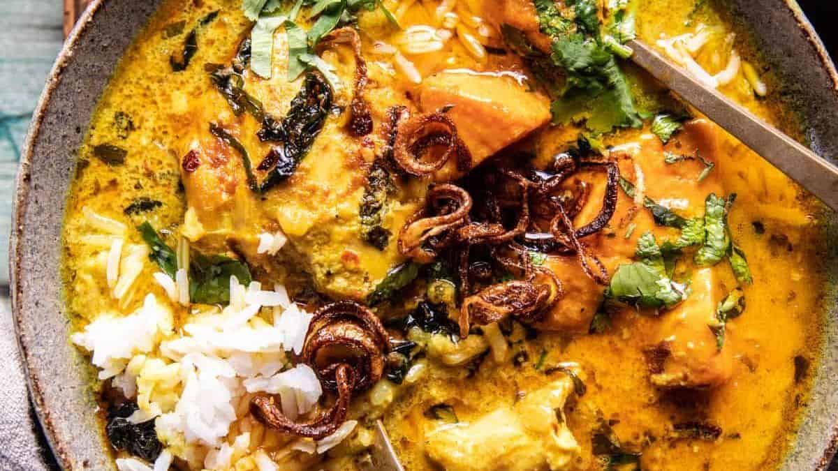A bowl of yellow curry with chicken chunks, white rice, fresh herbs, crispy fried onions, and a spoon.