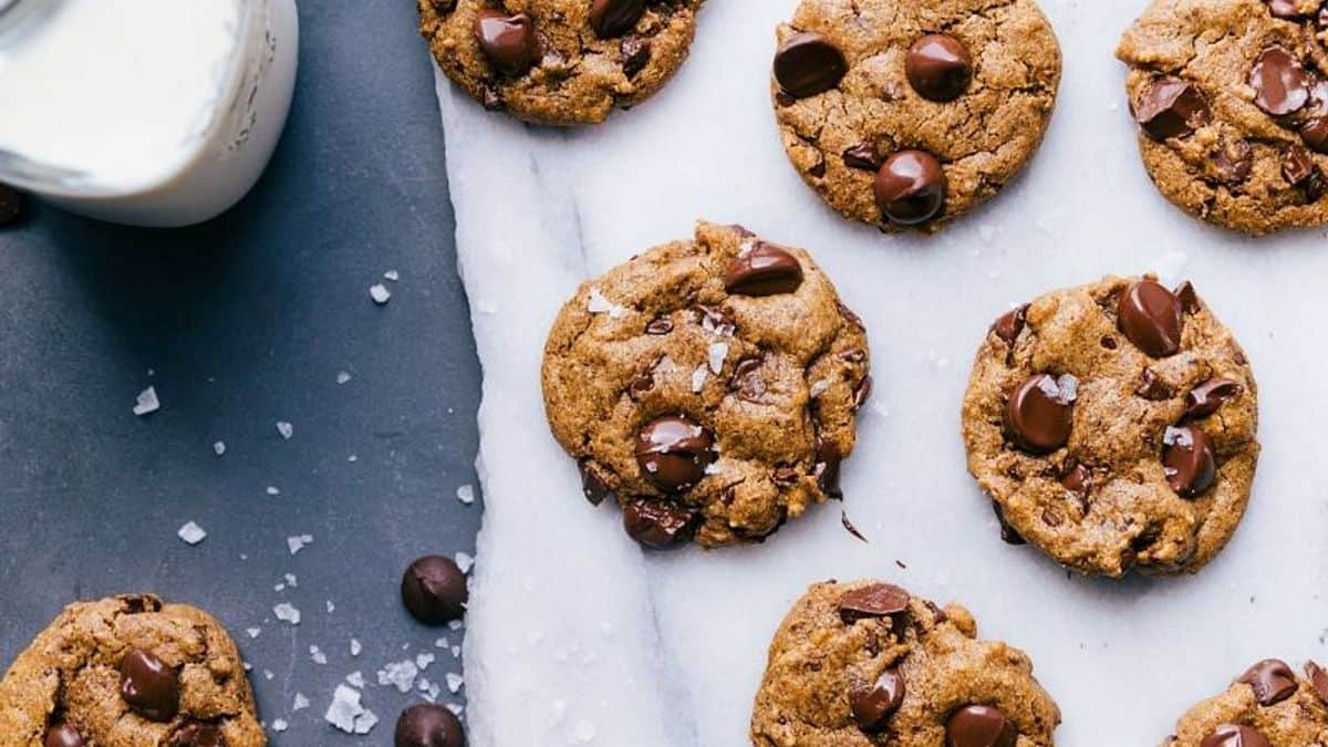 Chocolate chip cookies on a white marble surface beside a glass of milk, scattered chocolate chips, and a sprinkle of sea salt.
