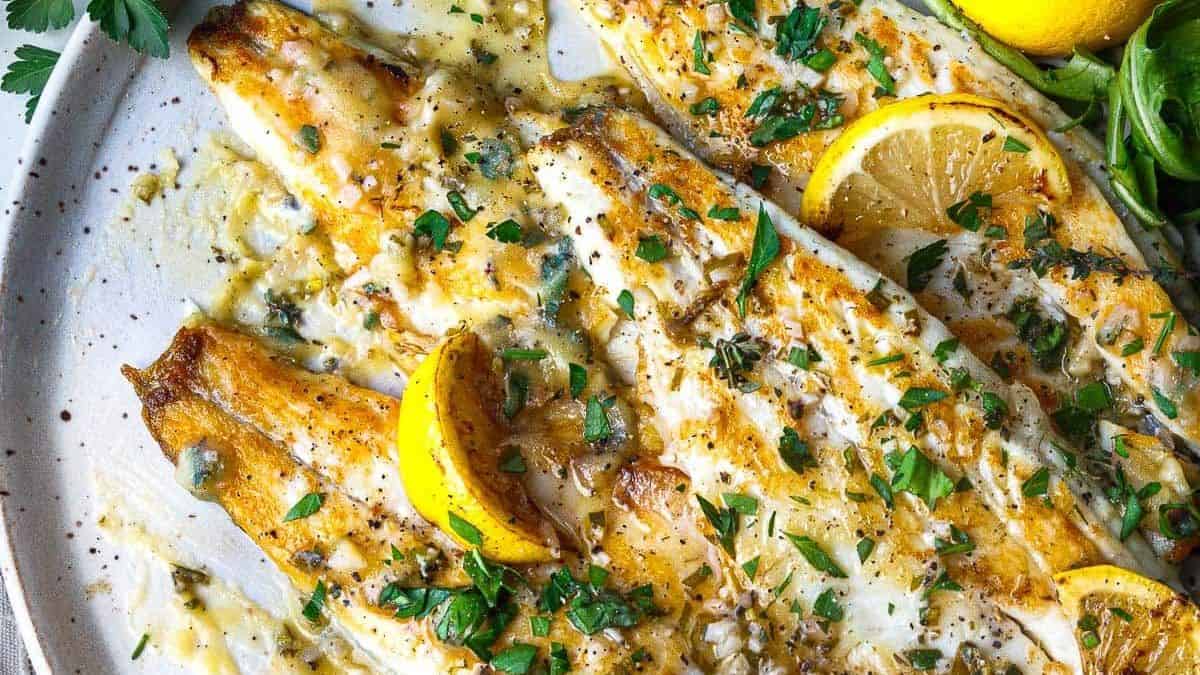 Cooked white fish fillets with lemon slices, chopped parsley, and creamy sauce on a white plate.
