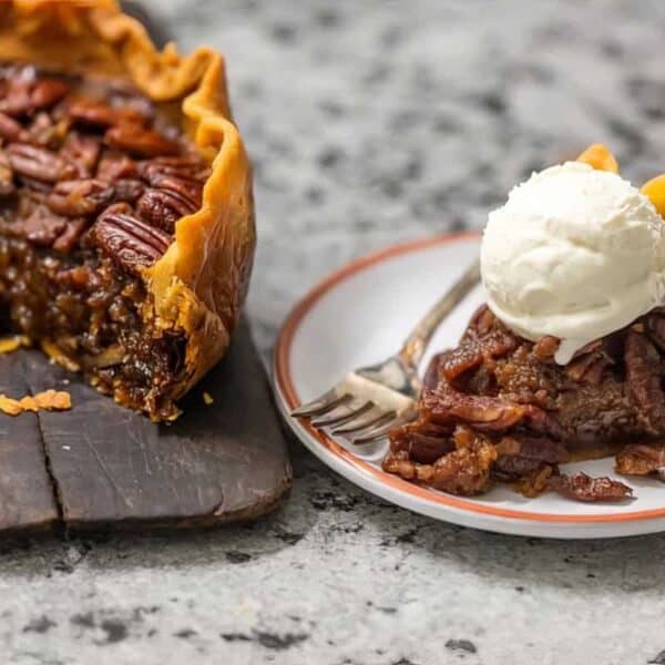 A sliced pecan pie sits on a cutting board, with one piece on a plate topped with a scoop of vanilla ice cream.