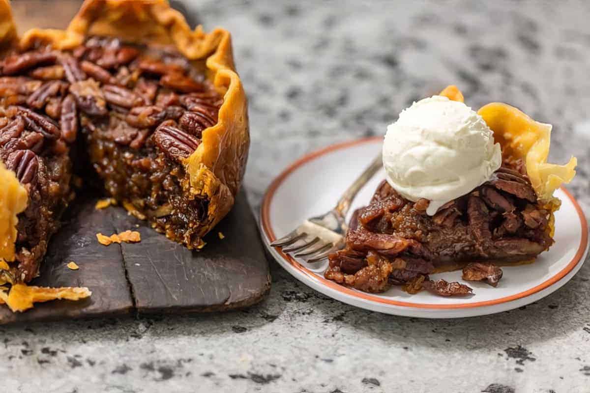 A sliced pecan pie sits on a cutting board, with one piece on a plate topped with a scoop of vanilla ice cream.