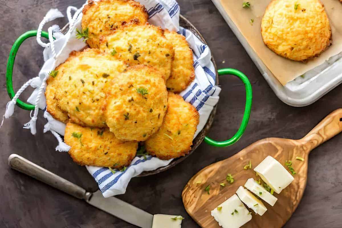 A cloth-lined basket with baked cheese biscuits, a sliced block of cheese, a knife, and one biscuit on a baking tray nearby.