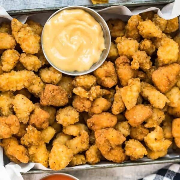 A tray lined with parchment paper holds crispy popcorn chicken pieces and a small metal cup of dipping sauce in the center.