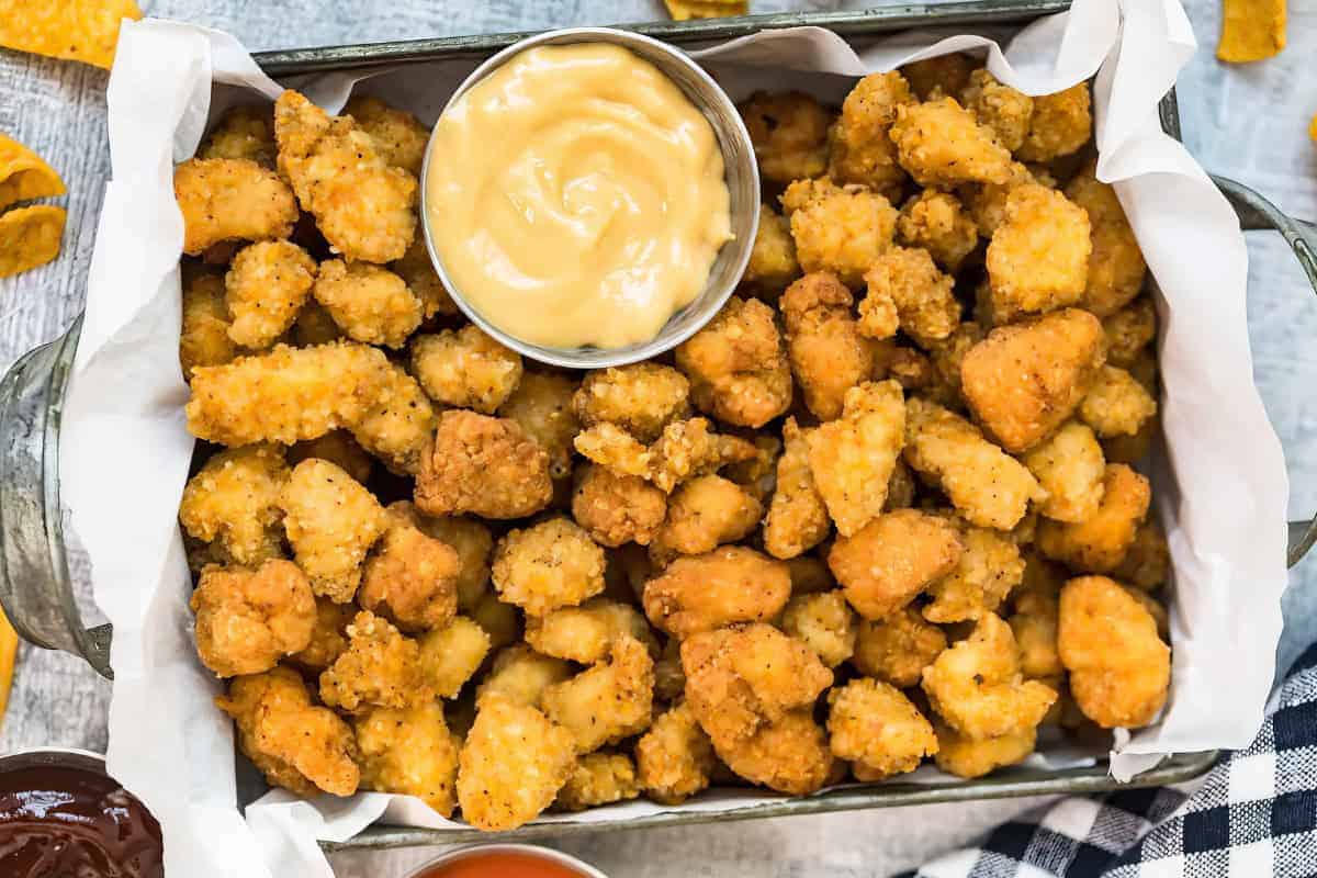 A tray lined with parchment paper holds crispy popcorn chicken pieces and a small metal cup of dipping sauce in the center.