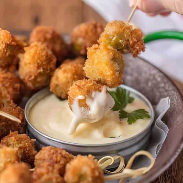 A hand dips a breaded olive skewer into a dish of creamy sauce; additional skewers rest on a nearby platter.