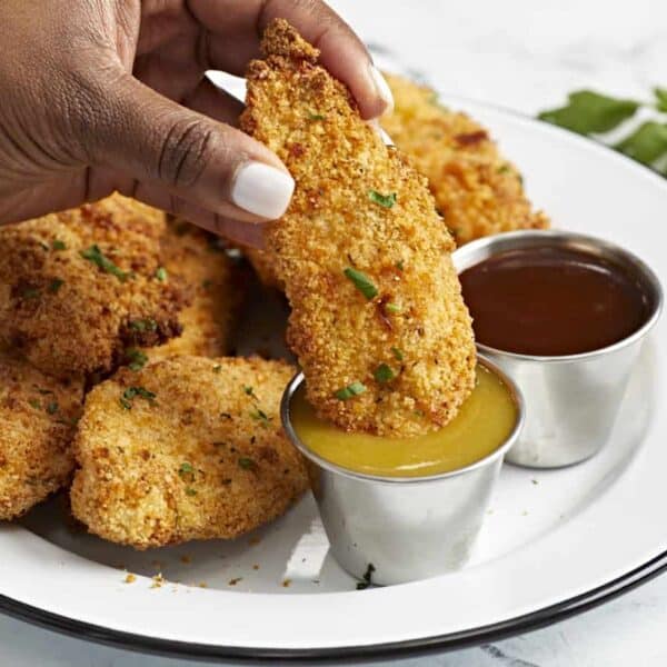 Breaded chicken strips, a cup of yellow sauce, a brown dipping sauce, and a white plate are shown.