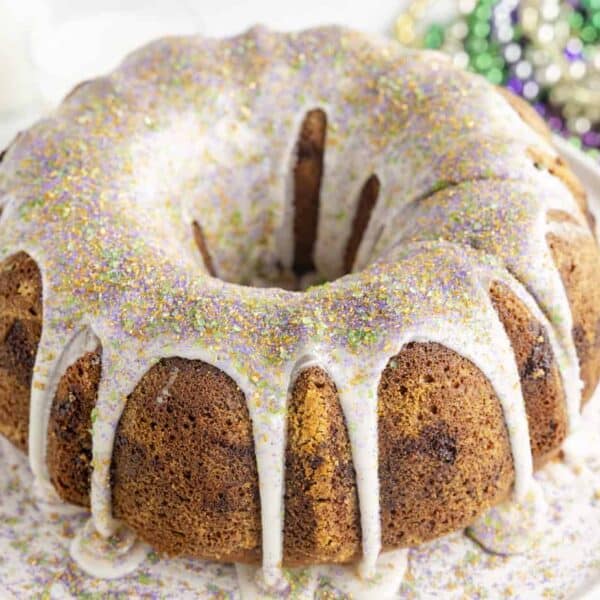 A bundt cake with white icing and purple, green, and gold sugar sprinkles sits on a plate. Mardi Gras beads are behind it.