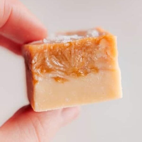 A hand holds a square piece of caramel fudge with a smooth, creamy texture and light brown color.