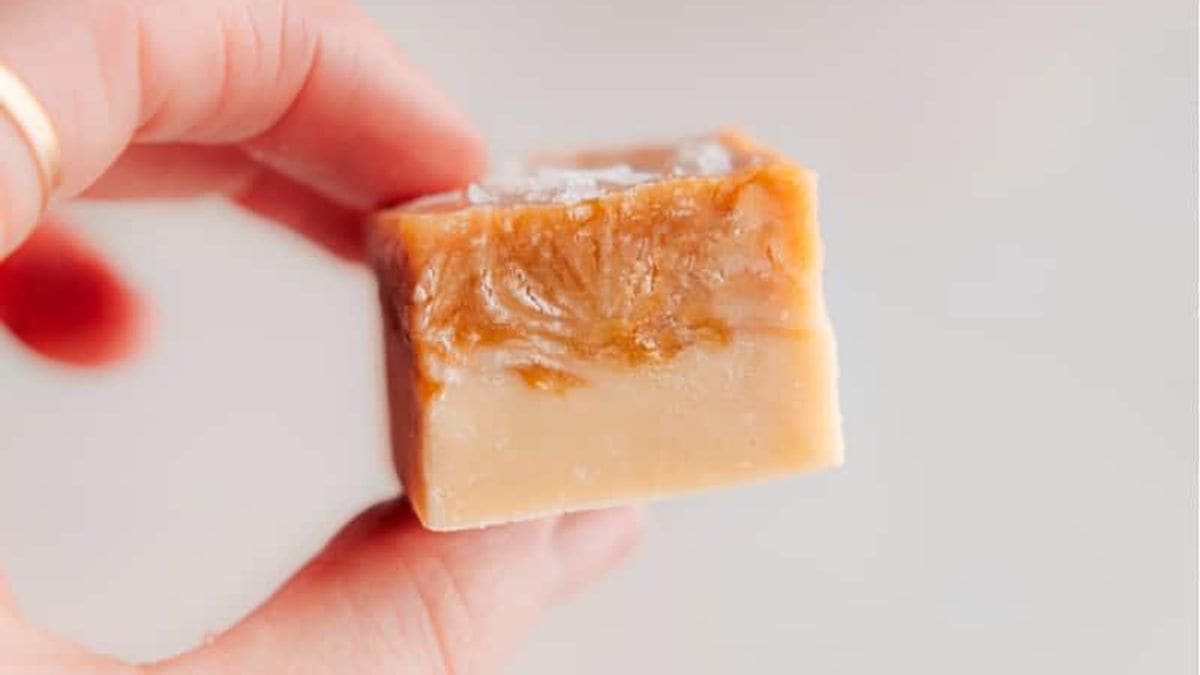 A hand holds a square piece of caramel fudge with a smooth, creamy texture and light brown color.