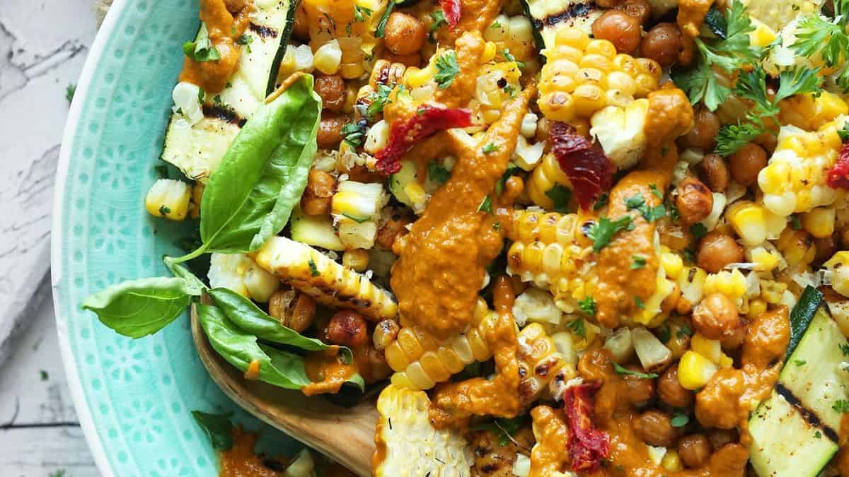 Grilled corn, chickpeas, zucchini, sun-dried tomatoes, basil, and parsley on a plate with creamy orange sauce on top.