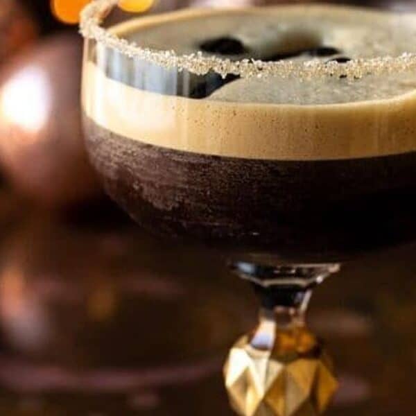 A glass of coffee cocktail with a frothy top and sugared rim sits on a reflective surface; blurred lights and ornaments in background.