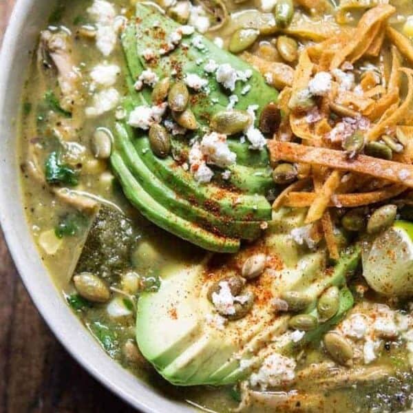 Green soup in a bowl with sliced avocado, tortilla strips, pumpkin seeds, corn, crumbled cheese, lime wedges, and spices.