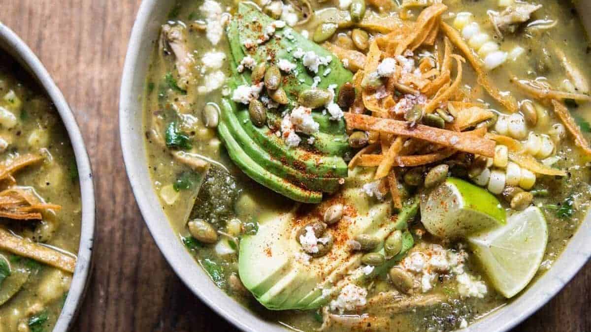 Green soup in a bowl with sliced avocado, tortilla strips, pumpkin seeds, corn, crumbled cheese, lime wedges, and spices.