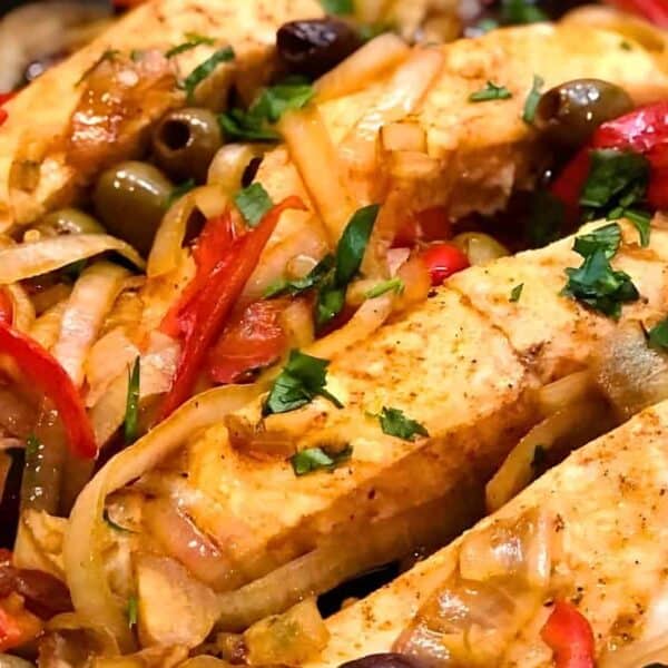 Chicken breasts in a skillet with sliced onions, red bell peppers, green olives, and scattered fresh herbs.