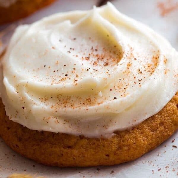 A frosted cookie with white icing, lightly sprinkled with cinnamon, sits on a parchment-lined surface next to several cinnamon sticks and dried wheat.