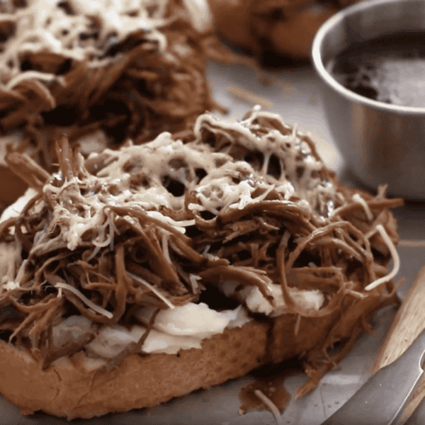 Toasted bread with shredded beef, melted cheese, spread, a metal cup of dipping sauce, and a fork on a tray.