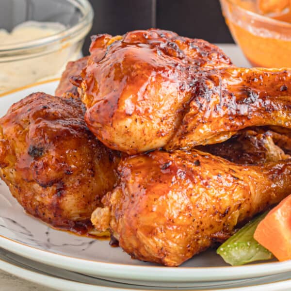 Barbecue-glazed chicken drumsticks on a plate with celery and carrot sticks, and bowls of dipping sauce in the background.