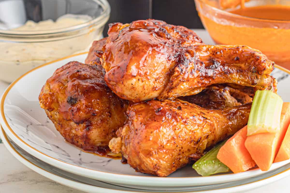 Barbecue-glazed chicken drumsticks on a plate with celery and carrot sticks, and bowls of dipping sauce in the background.