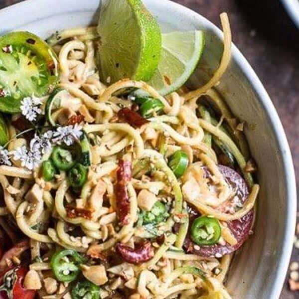 Noodle salad in a bowl with cherry tomatoes, green chilies, lime wedges, chopped peanuts, herbs, and a bowl of peanuts behind.