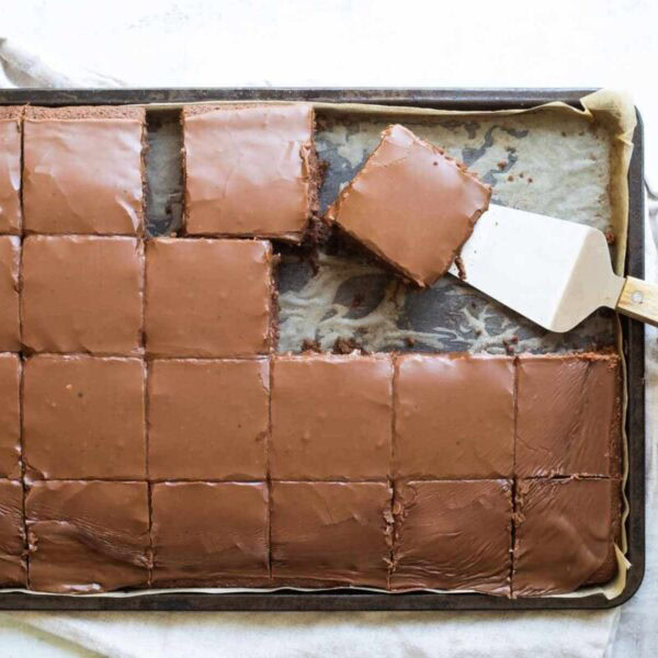 A tray of chocolate-frosted sheet cake cut into squares, with one piece being lifted out with a spatula.
