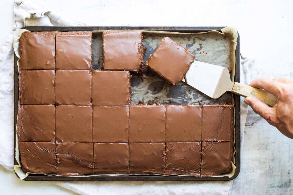 A hand uses a spatula to lift a square piece of chocolate sheet cake from a parchment-lined baking tray with several pieces left.