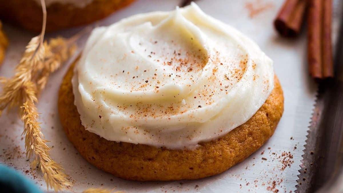 A thick, round cookie with creamy white frosting on top is lightly dusted with cinnamon. It sits on a baking sheet near a piece of wheat and several cinnamon sticks.