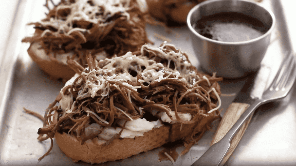 Toasted bread with shredded beef, melted cheese, spread, a metal cup of dipping sauce, and a fork on a tray.