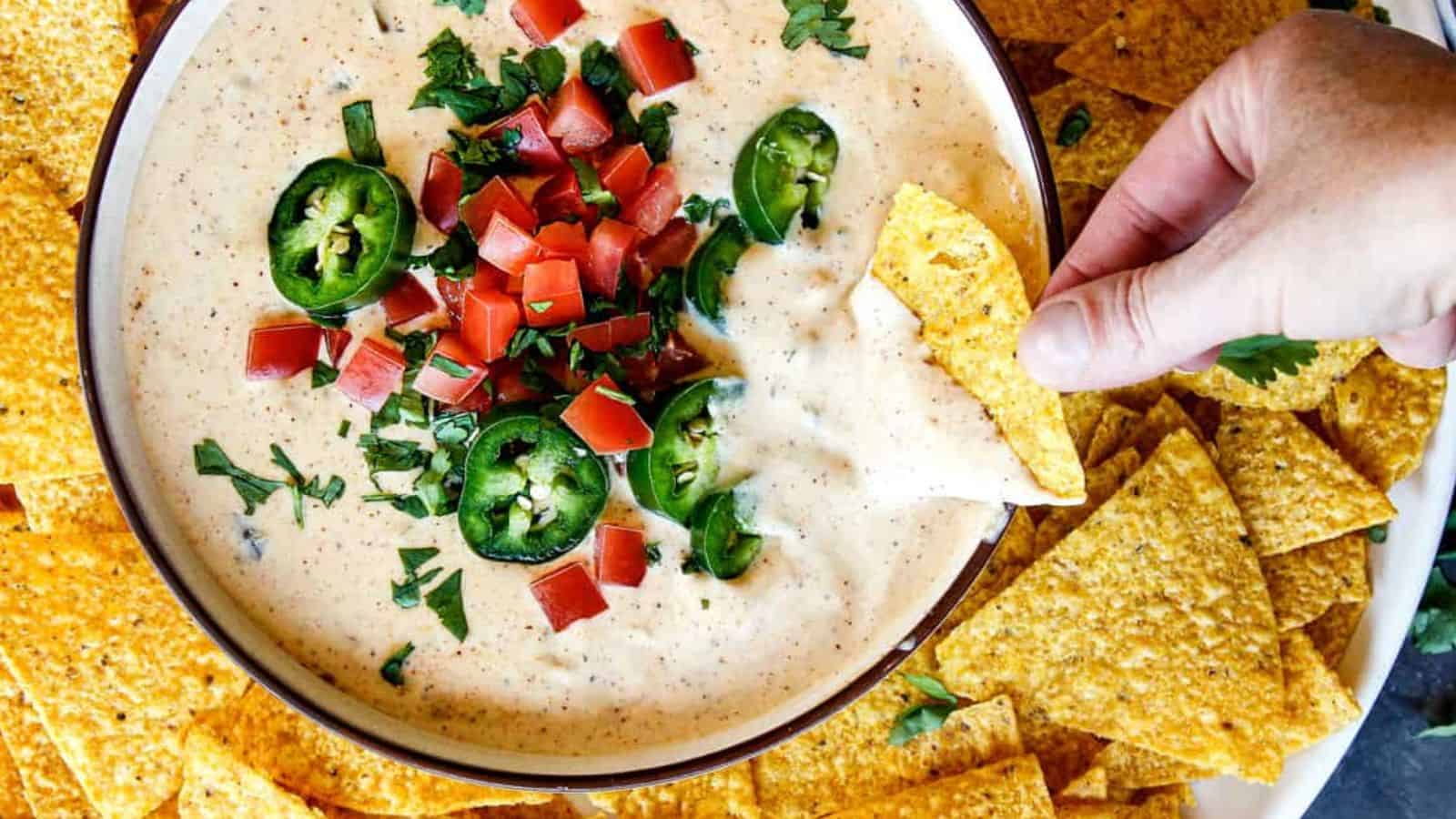 A hand dips a tortilla chip into queso dip topped with diced tomatoes, jalapeños, and chopped cilantro, surrounded by chips.