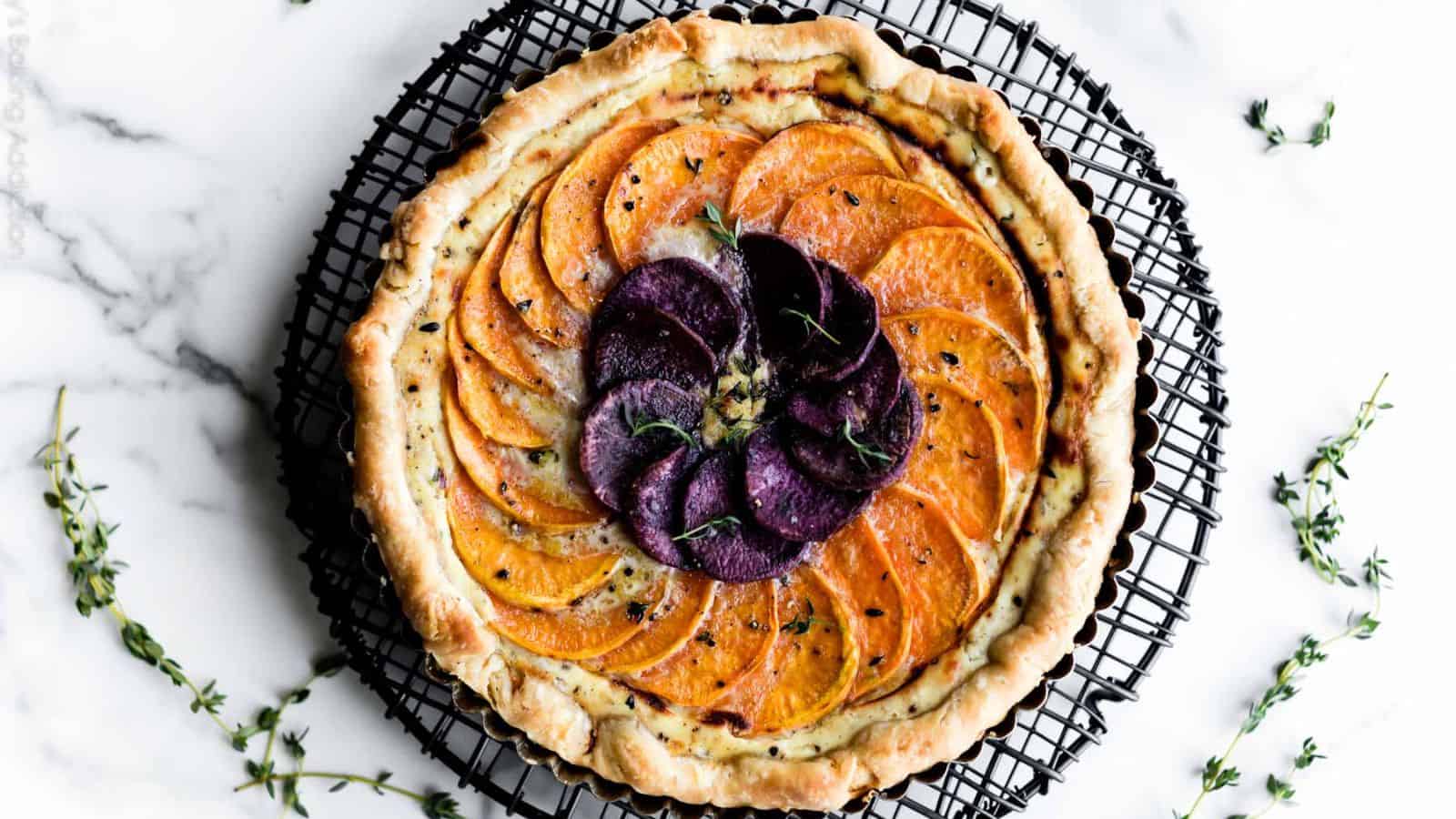A tart with a flaky crust topped with overlapping orange and purple sweet potato slices on a black wire cooling rack.