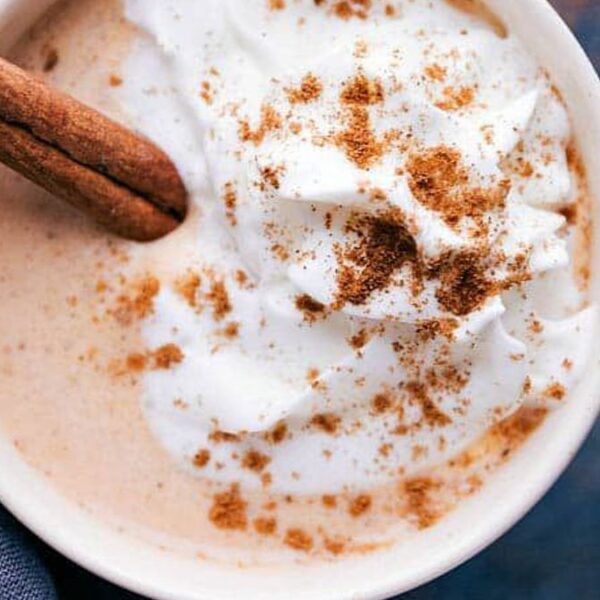 A mug of creamy beverage topped with whipped cream, cinnamon sprinkles, and a cinnamon stick garnish.