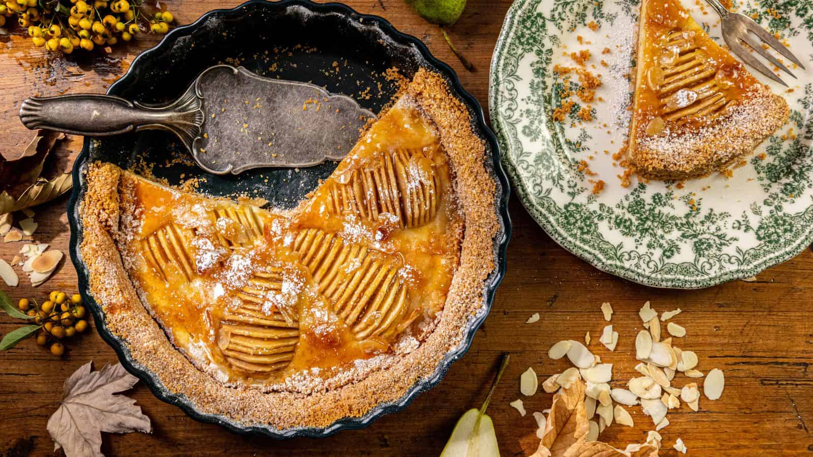 A pear tart in a fluted pie dish, a plate with one slice, scattered almonds, and autumn leaves on a wooden table.
