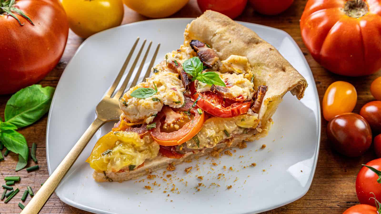 A slice of tomato pie topped with basil on a square white plate with a fork, surrounded by whole tomatoes and herbs.
