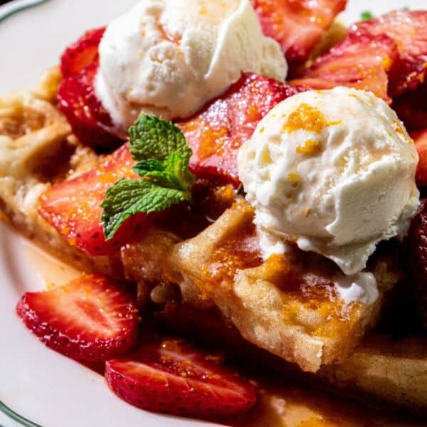Waffles on a plate with sliced strawberries, two scoops of vanilla ice cream, and mint leaves on top.