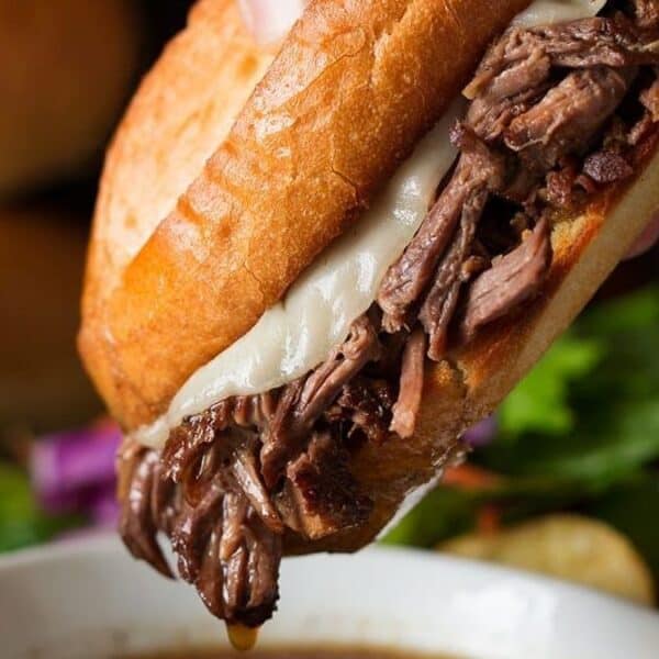 A hand holding a French dip sandwich with shredded beef and melted cheese, dipping the bread into a bowl of au jus.