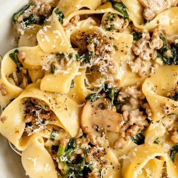 A plate of pappardelle pasta with creamy sauce, mushrooms, spinach, ground meat, and grated cheese, topped with black pepper.
