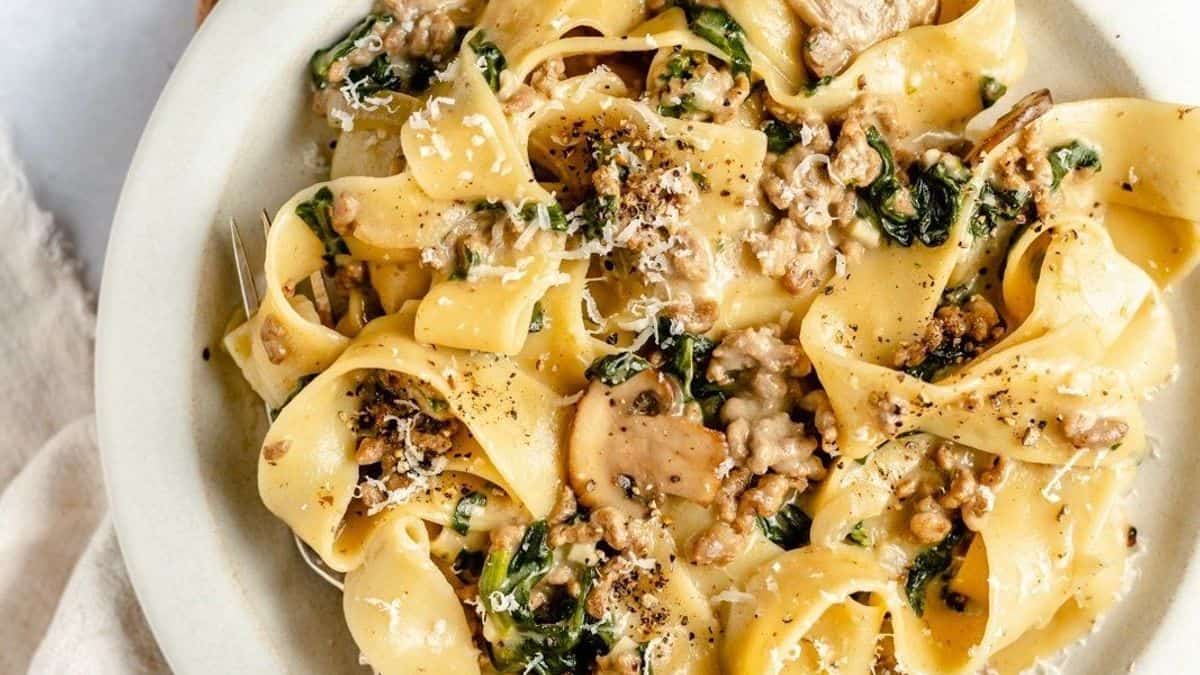 A plate of pappardelle pasta with creamy sauce, mushrooms, spinach, ground meat, and grated cheese, topped with black pepper.