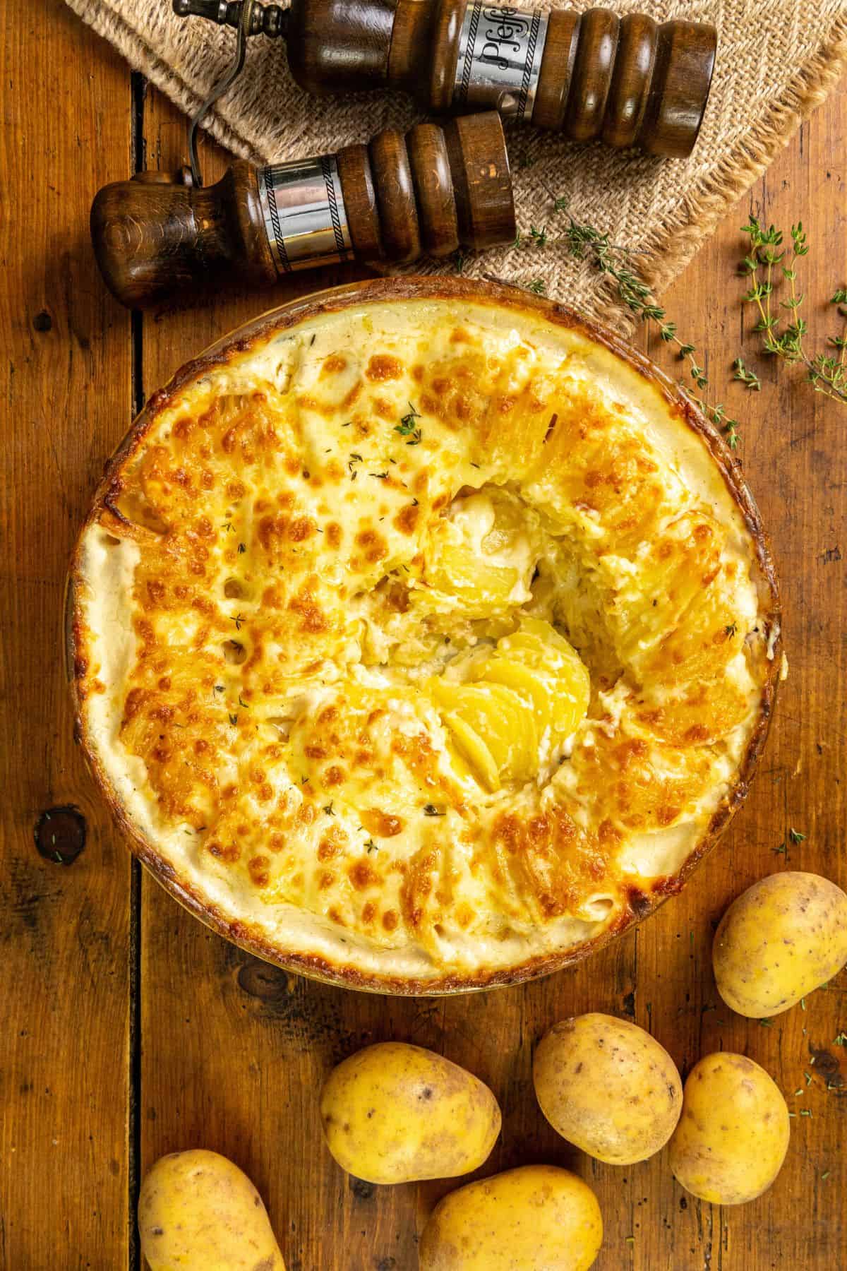 A baked gratin dauphinois with a golden, cheesy top, partly served, next to raw potatoes and pepper mills on a wooden table.