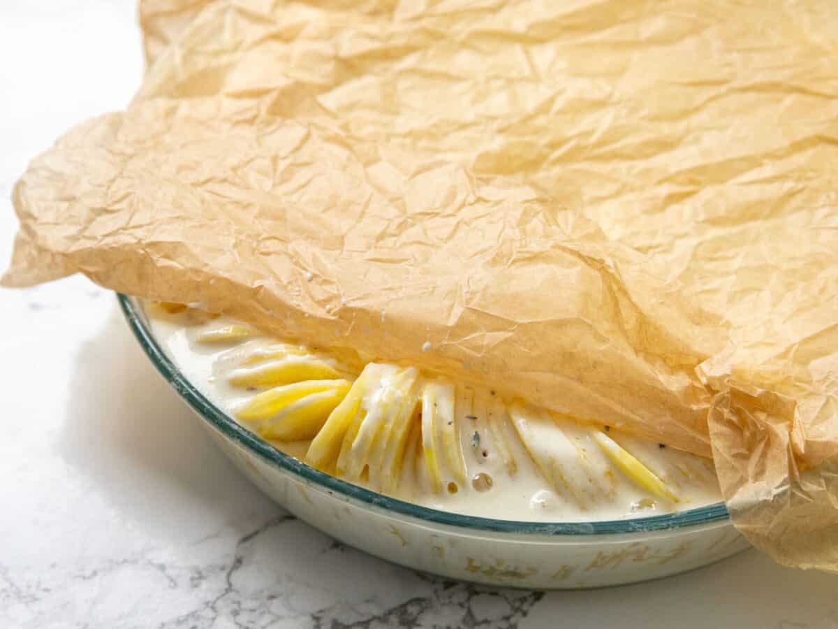 Glass dish with sliced potatoes in creamy sauce, partially covered by brown parchment paper.
