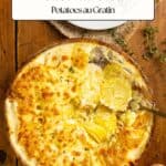 A baking dish of golden-brown gratin dauphinois on a wooden table with a serving spoon and whole potatoes nearby.