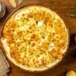 A round dish of gratin dauphinois on a wooden table with raw potatoes, fresh herbs, a jar of bay leaves, and a woven mat.